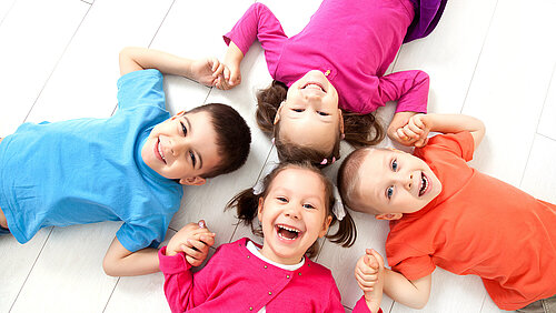 Startbild: Foto von fröhlichen Kindern, die im Kreis auf dem Boden liegen und sich an den Händen fassen