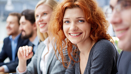 Startbild: Foto von Menschen bei einem Vortrag, im Vordergrund eine rothaarige Frau, die in die Kamera lächelt
