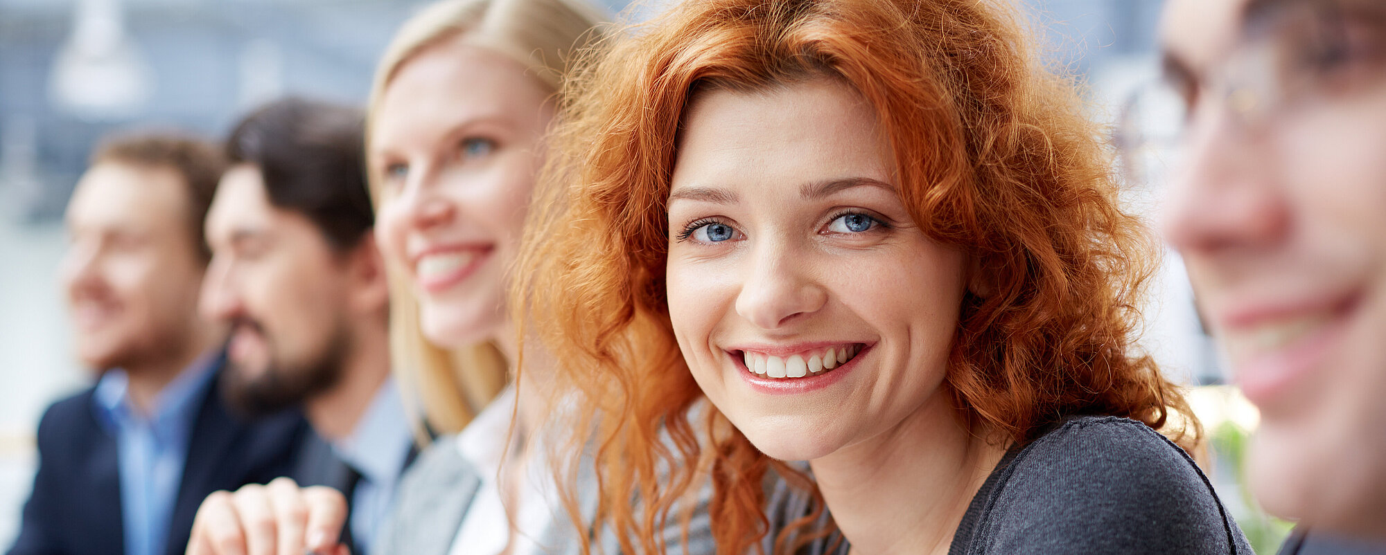 Startbild: Foto von Menschen bei einem Vortrag, im Vordergrund eine rothaarige Frau, die in die Kamera lächelt