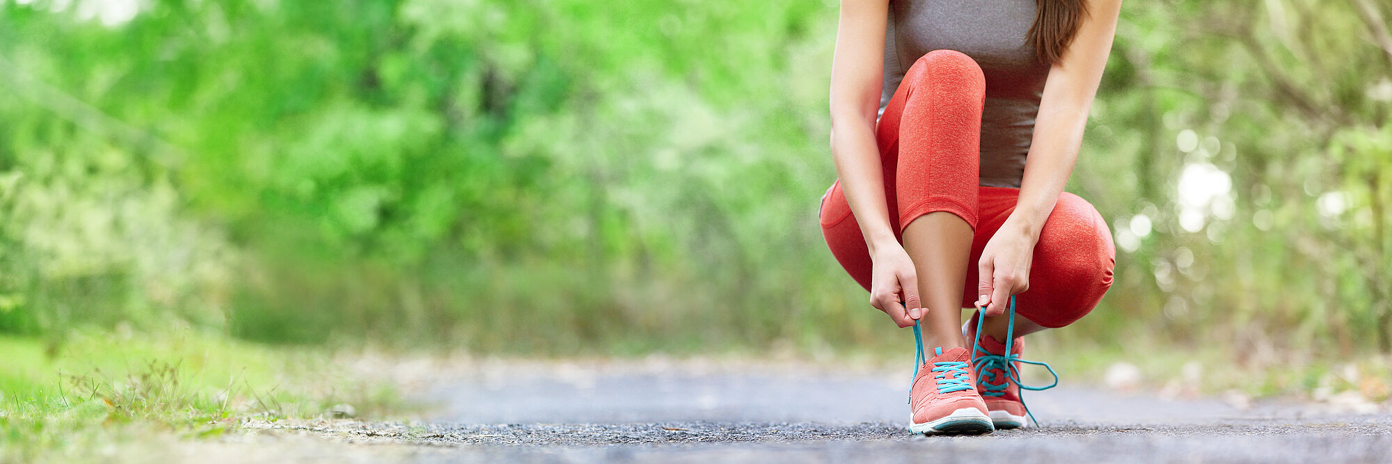 Startbild: Foto einer jungen Frau, die Sportkleidung trägt und auf einem Waldweg ihre Sportschuhe bindet