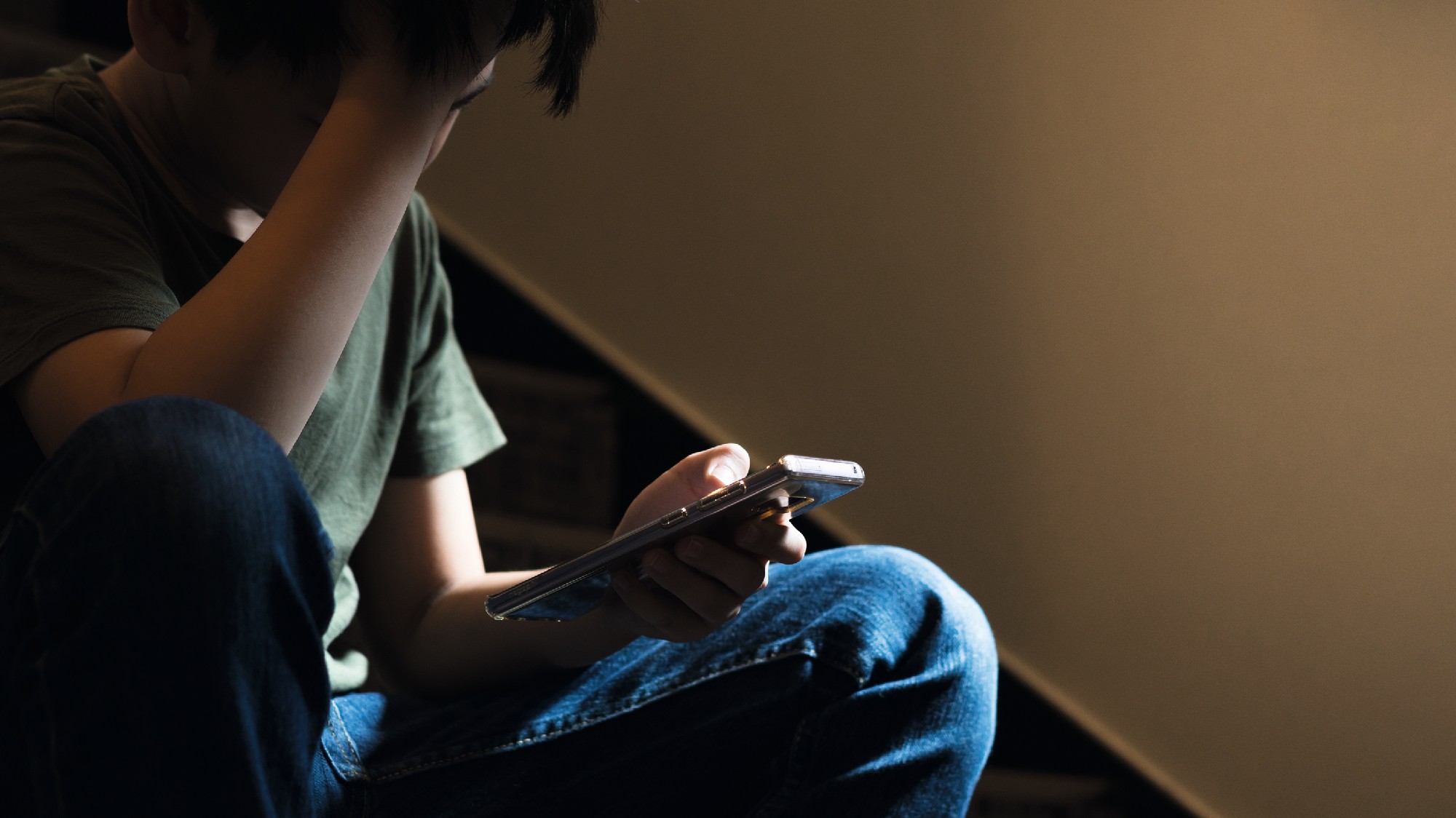 Ein junge sitzt in einem dunklen Treppenhaus auf einer Treppe, blickt auf ein Smartphone in der linken Hand und stützt den Kopf mit der rechten Hand