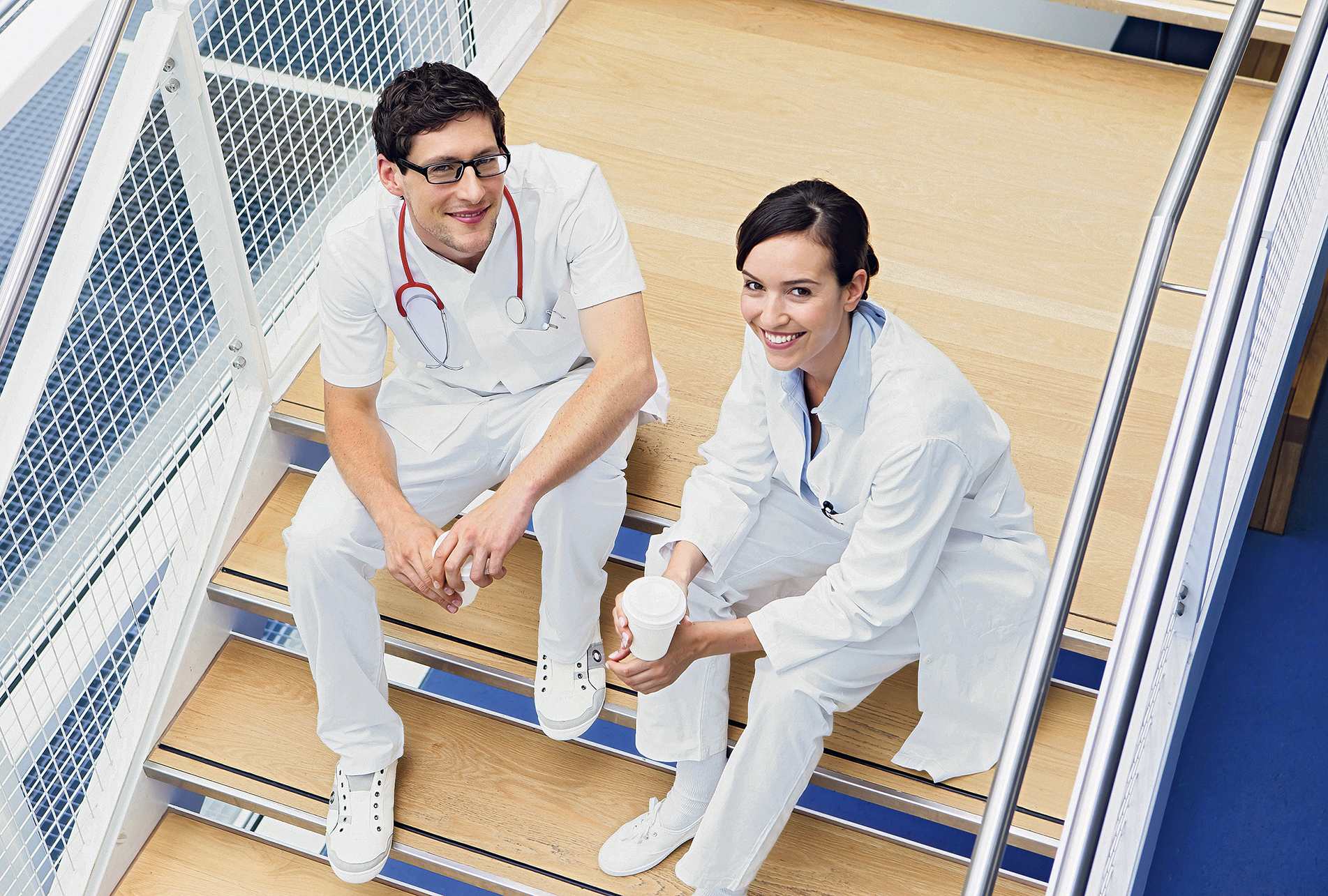 Startbild: Foto von einem jungen Arzt und einer jugen Ärztin, die auf der Treppe eines Krankenhauses sitzen