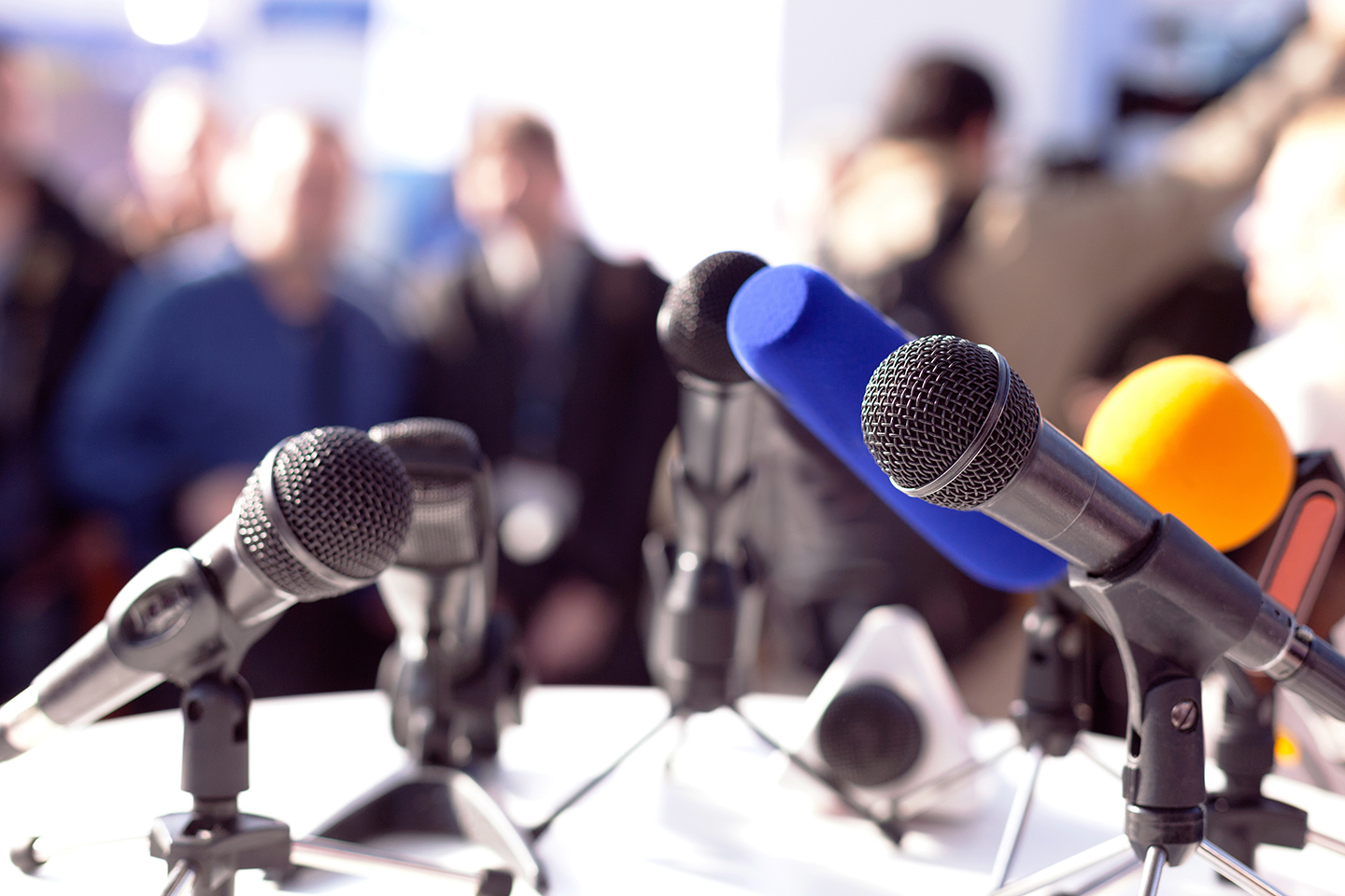 Startbild: Foto von mehreren Mikrofonen im Vordergrund, im Hintergrund unscharf Journalisten bei einer Pressekonferenz